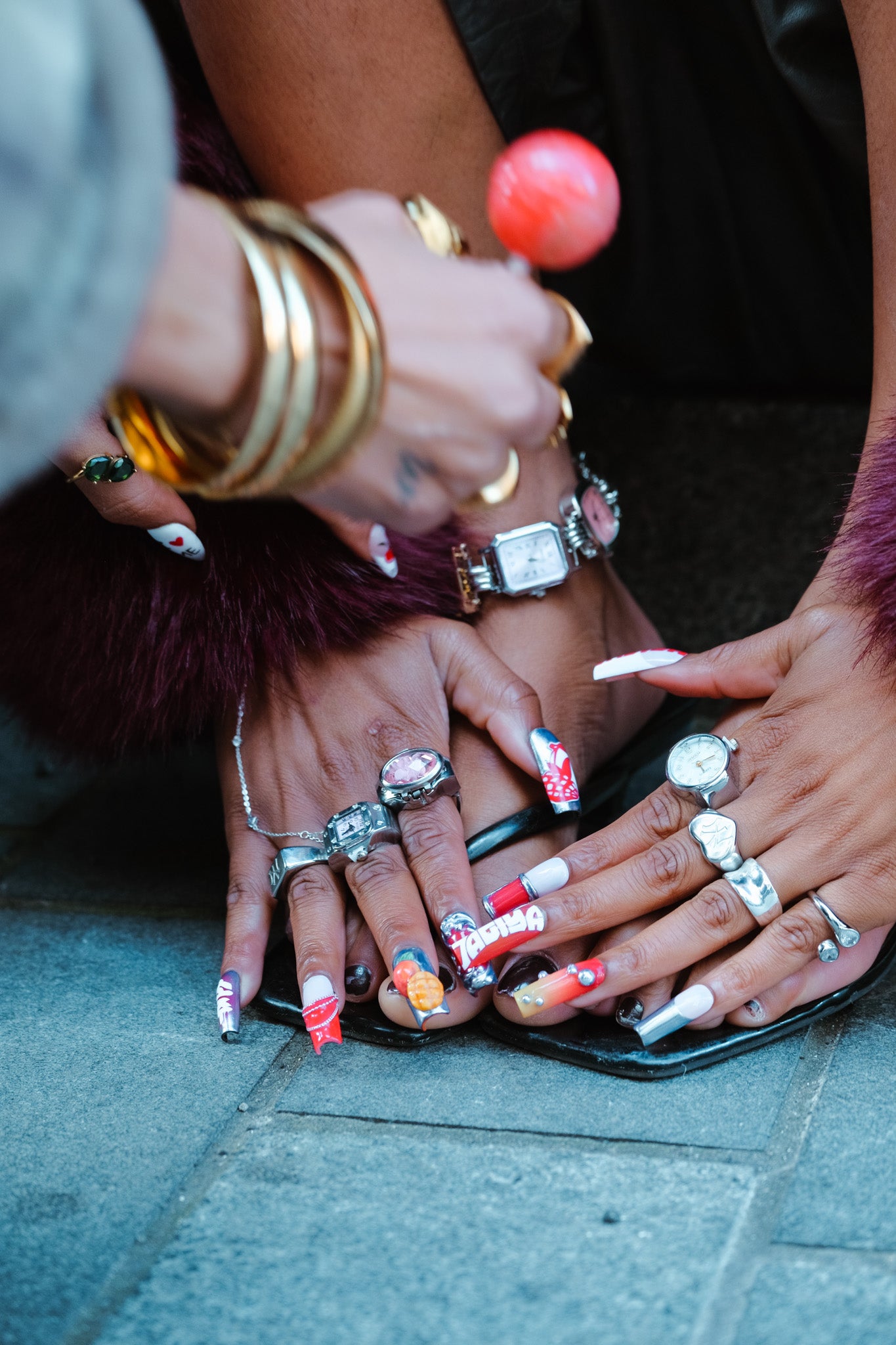 Close-up of hands with colorful nail polish and multiple rings on a pavement background