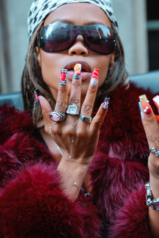 Person wearing sunglasses and a red fur coat with colorful nail polish and multiple rings.
